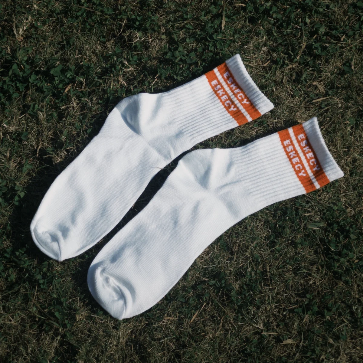 Eskecy white crew socks with orange logo stripe, laid flat outdoors on grass